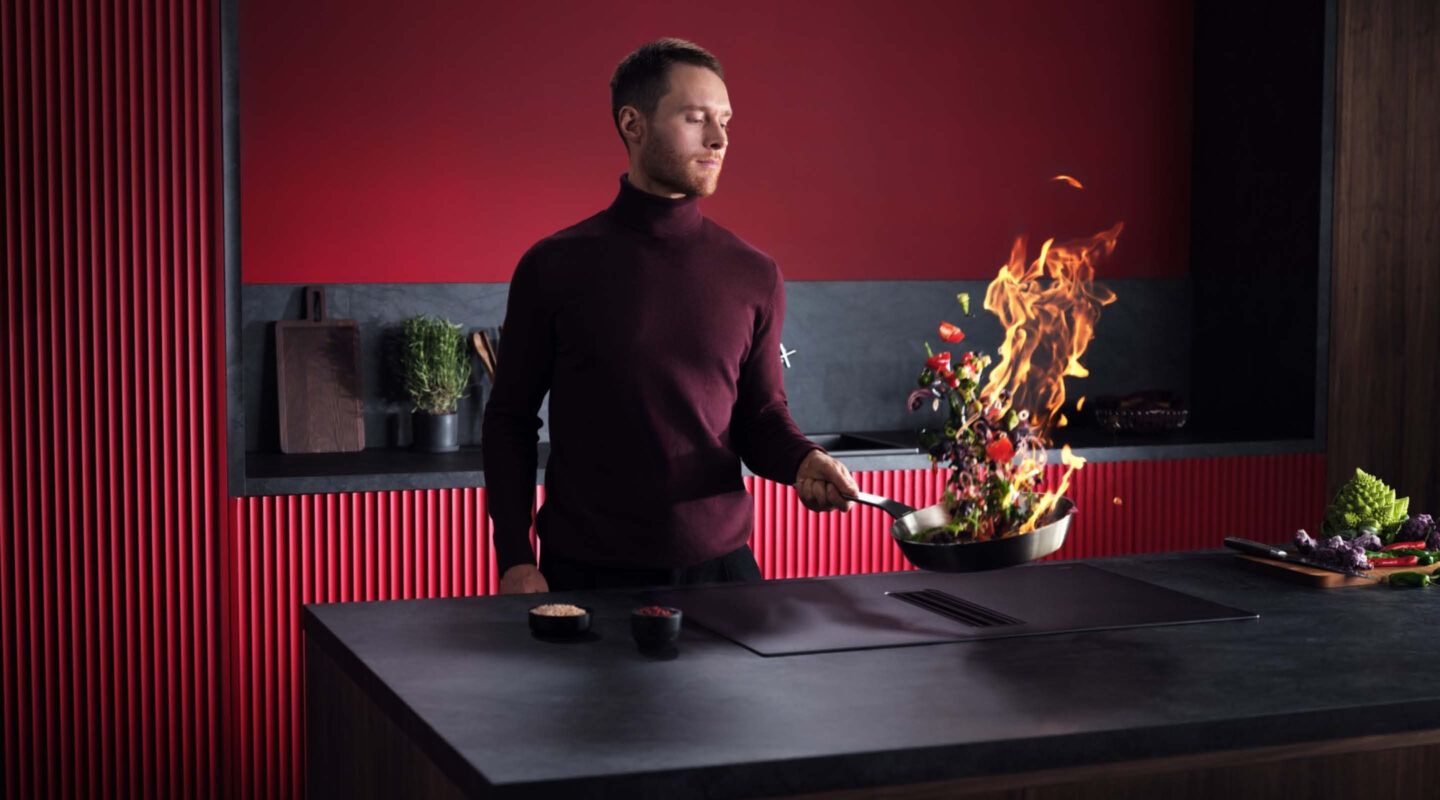 Ein Mann in einem roten Rollkragenpullover schwänkt eine Pfanne aus dem gebratenes Gemüse und Flammen aufsteigen.