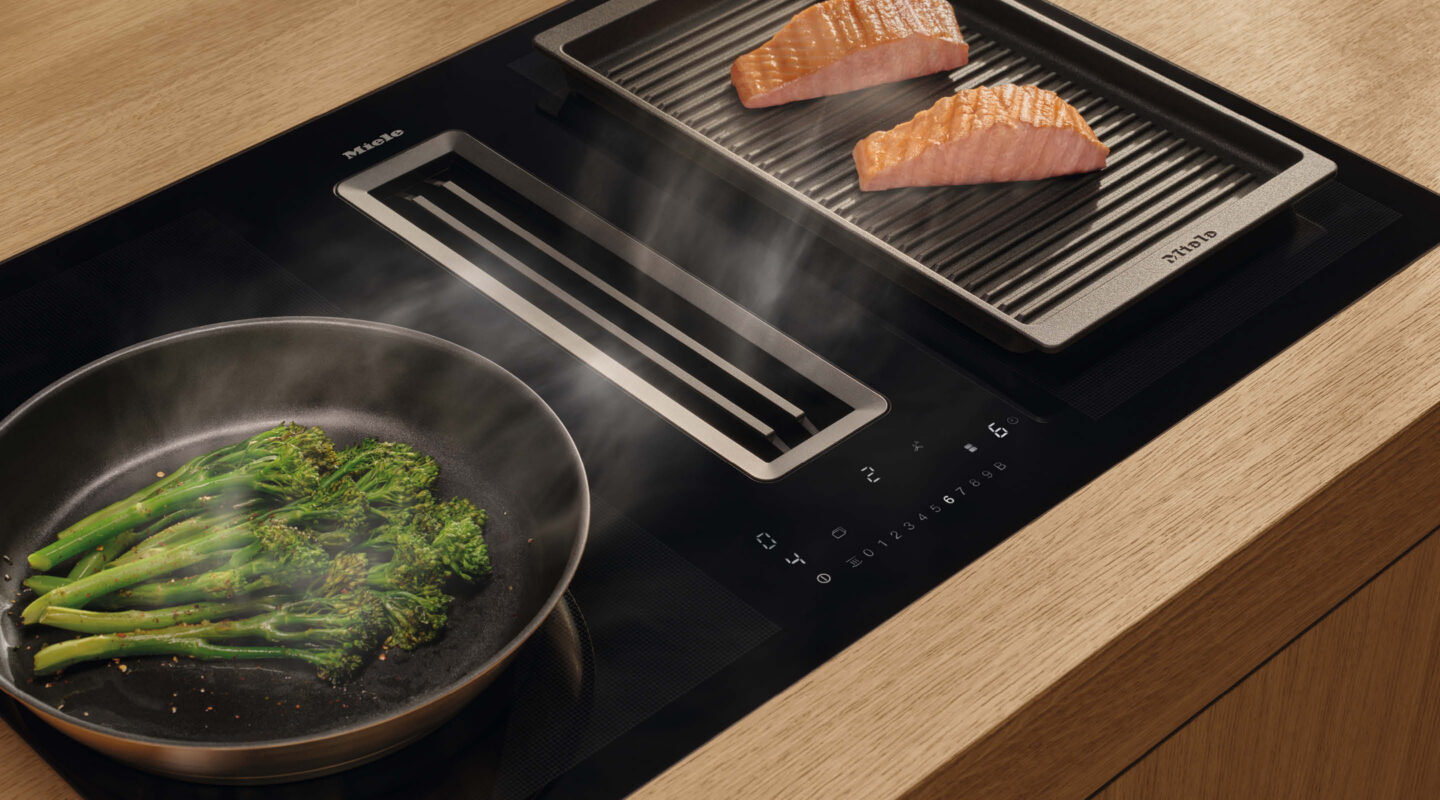 Induktionskochfeld von Miele mit integriertem Dunstabzug, Grillplatte mit Lachs und Pfanne mit Brokkoli auf einer hellen Holzarbeitsplatte.