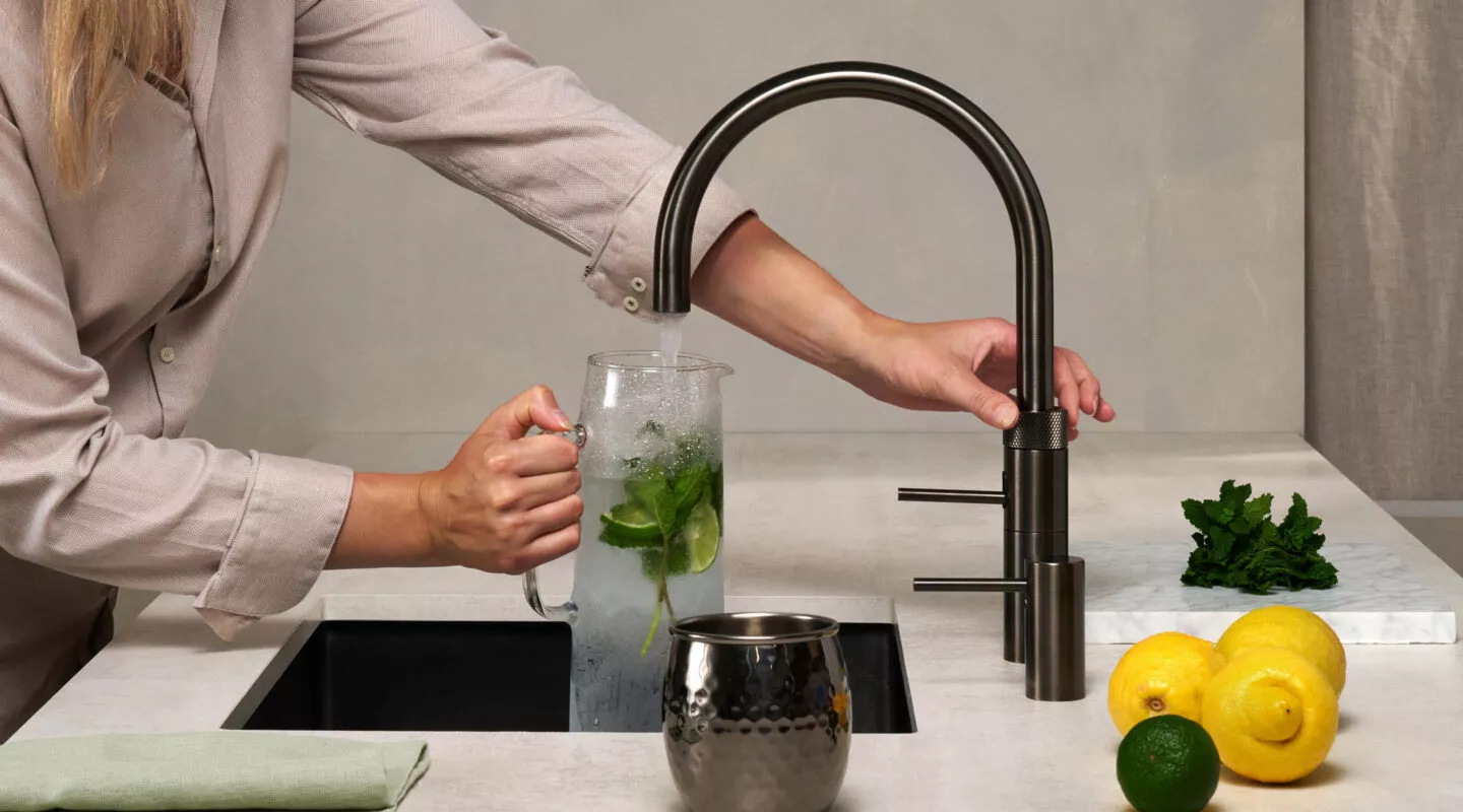 Ein schwarzer runder Kochendwasserhahn von Quooker. Eine Frau befüllt eine Karaffe mit Wasser.