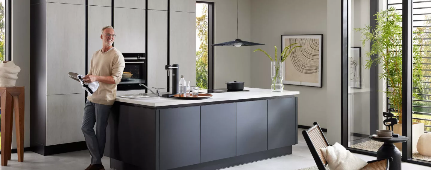 Moderne Küche mit Kochinsel in dunklem Mattdesign und heller Arbeitsplatte, ein Mann steht entspannt am Tresen – stilvolles, lichtdurchflutetes Wohnambiente mit klaren Linien
