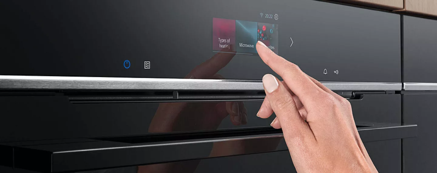 Detailaufnahme eines modernen Siemens Backofens mit hochauflösendem Touchdisplay, auf dem eine Hand verschiedene Kochfunktionen auswählt, integriert in eine elegante Küchenfront
