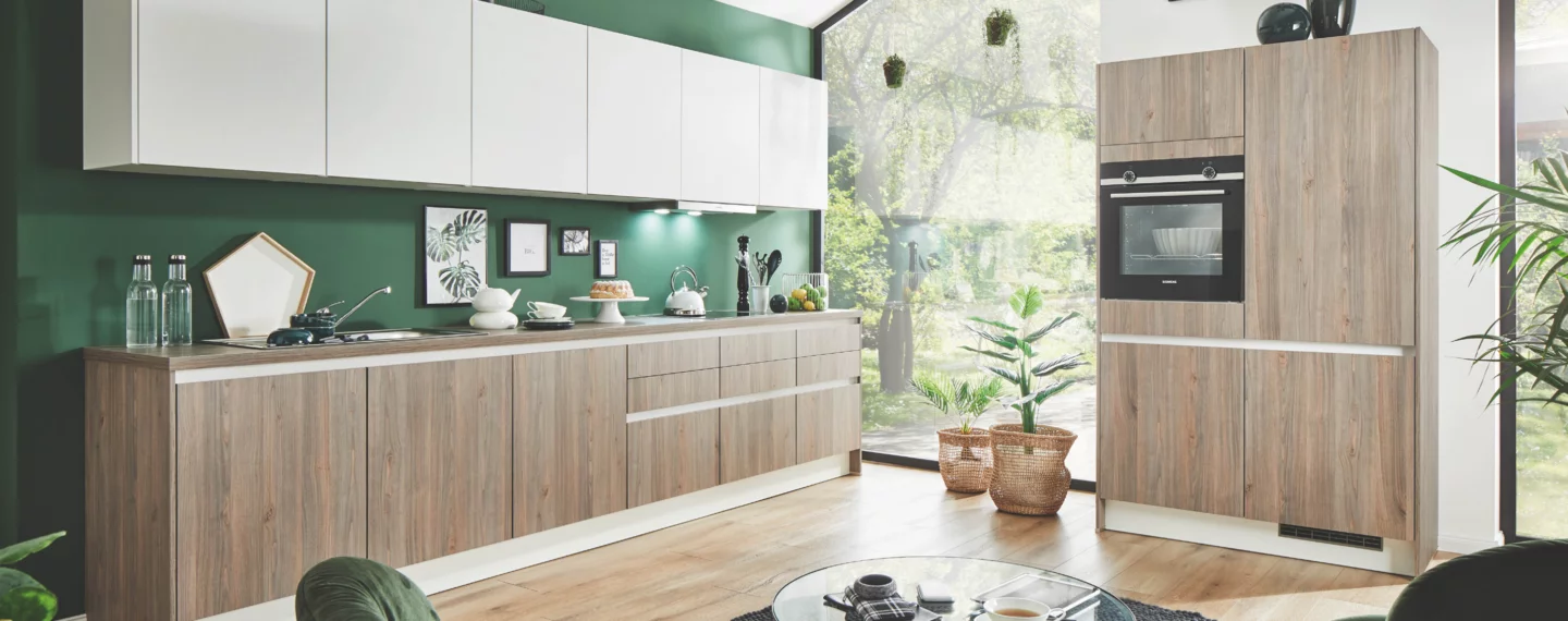 Moderne Küchenplanung mit griffloser Einbauküche in heller Holzoptik, weißen Oberschränken und grüner Rückwand, Spüle und Kochbereich sowie Hochschrank mit Siemens-Backofen und integriertem Kühlschrank – hochwertige Küchenmöbel in hellem, wohnlichem Ambiente.