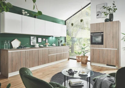 Moderne offene Wohnküche mit grifflosen Küchenmöbeln in heller Holzoptik, weißen Oberschränken und dunkelgrüner Rückwand, ergänzt durch Hochschrank mit Einbaubackofen, große Fenster und einen wohnlichen Sitzbereich – stilvolle individuelle Küchenplanung.