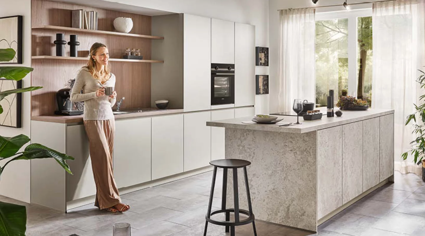 Helle moderne Wohnküche mit großer Kücheninsel, grifflosen weißen Küchenmöbeln, Holzelementen und integriertem Einbaubackofen; eine Frau mit Tasse steht an der Küchenzeile, während große Fenster und Pflanzen das offene Küchenkonzept ergänzen.