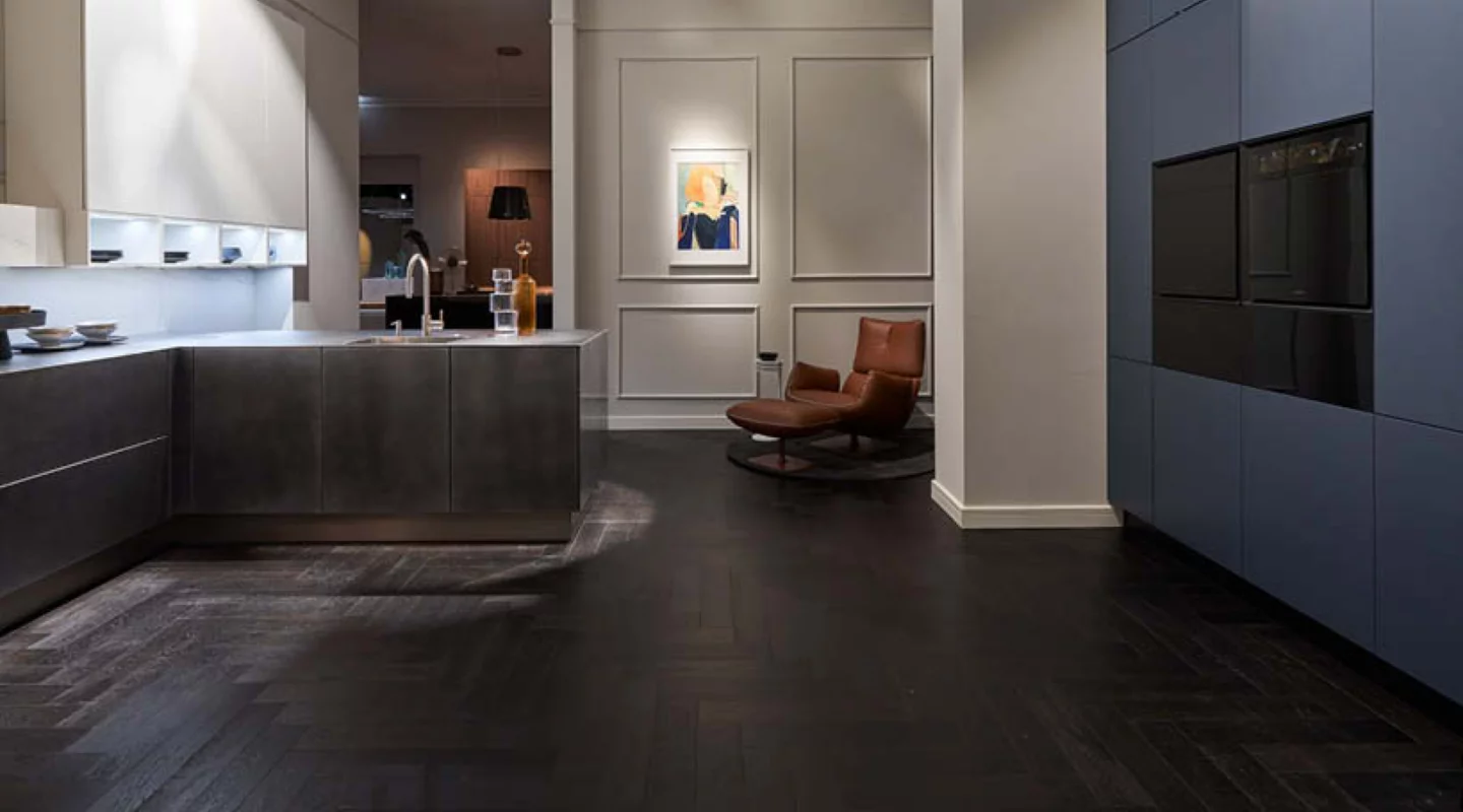 Moderne Designküche mit dunkler Kücheninsel und Spüle, weißen Schränken sowie grifflosen dunkelblauen Küchenmöbeln mit schwarzen Einbaugeräten; dunkler Holzboden und brauner Loungesessel verbinden Küche und Wohnbereich.