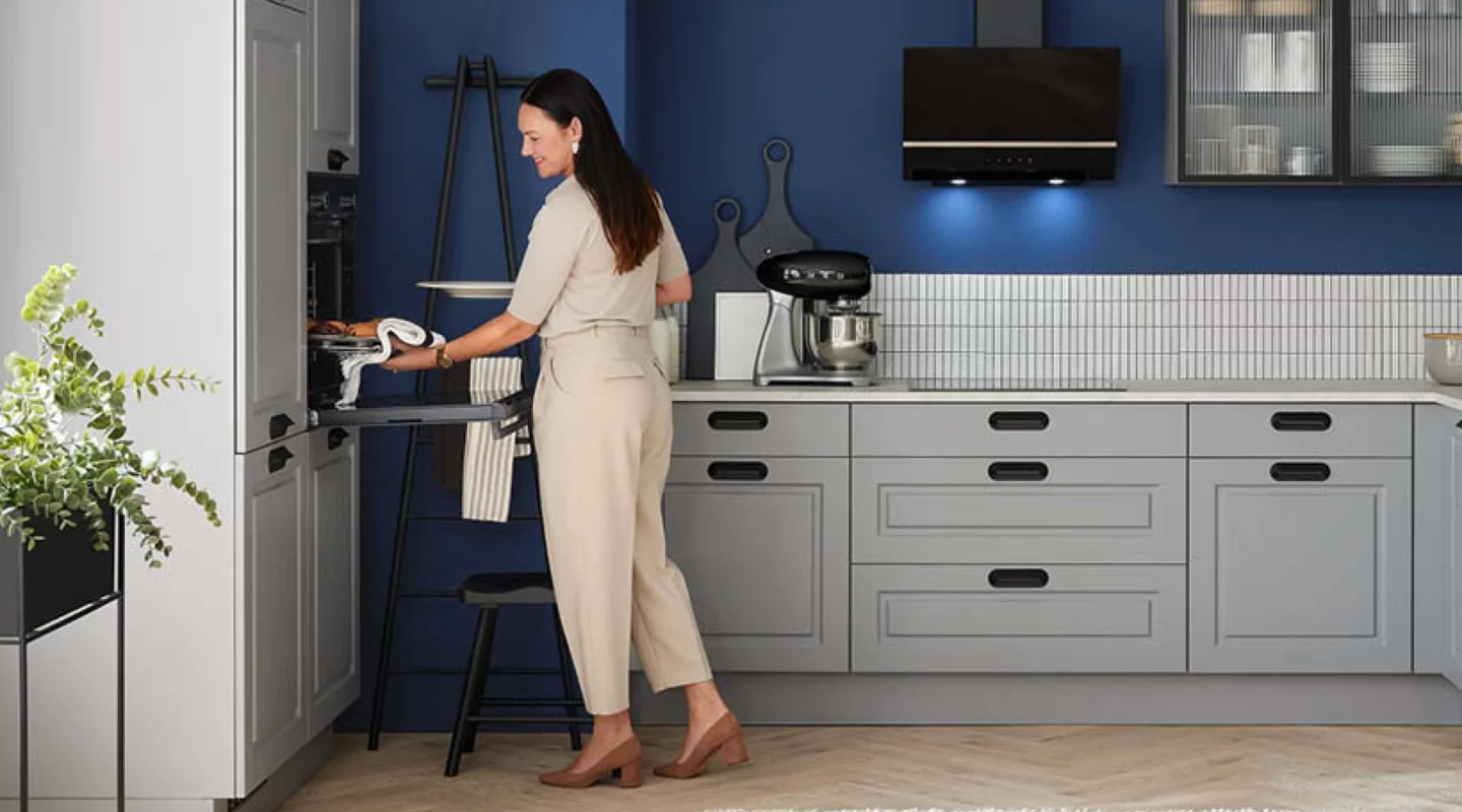 Moderne graue Landhausküche mit Rahmenfronten, schwarzer Dunstabzugshaube und Einbaubackofen vor dunkelblauer Wand; eine Frau entnimmt ein Backblech aus dem ergonomisch integrierten Ofen, während hochwertige Küchenmöbel und eine helle Arbeitsfläche das Küchendesign prägen.