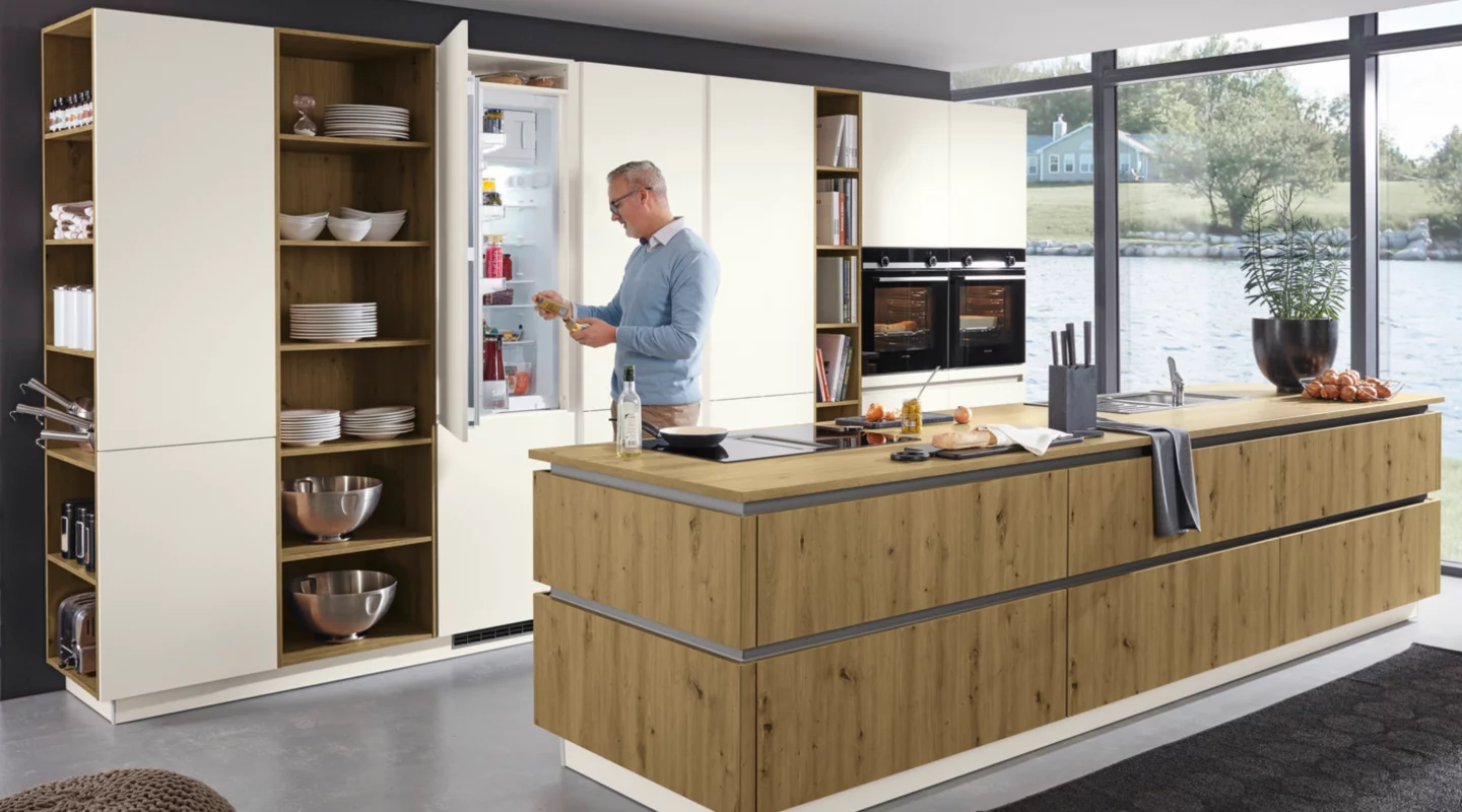 Moderne Wohnküche mit großer Kücheninsel in Holzoptik, grifflosen Küchenmöbeln, offenem Einbaukühlschrank und zwei Einbaubacköfen; ein Mann steht am Kühlschrank, während offene Regale, Kochfeld, Spüle und große Fenster die hochwertige Küchenplanung ergänzen.