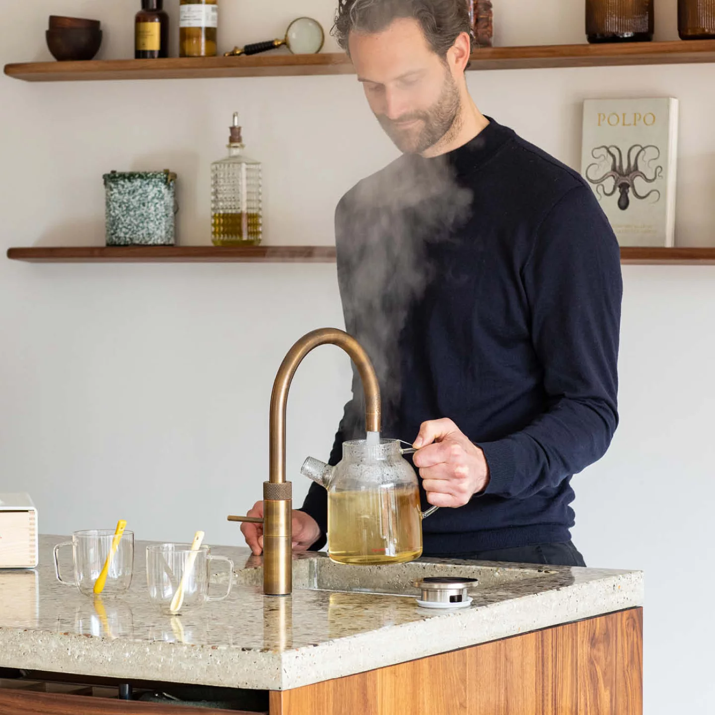 Mann füllt in einer modernen Küche eine Glaskanne mit heißem Wasser an einer bronzefarbenen Heißwasserarmatur; Kücheninsel mit Holzfronten, heller Arbeitsplatte und offenen Holzregalen ergänzt das hochwertige Küchendesign.