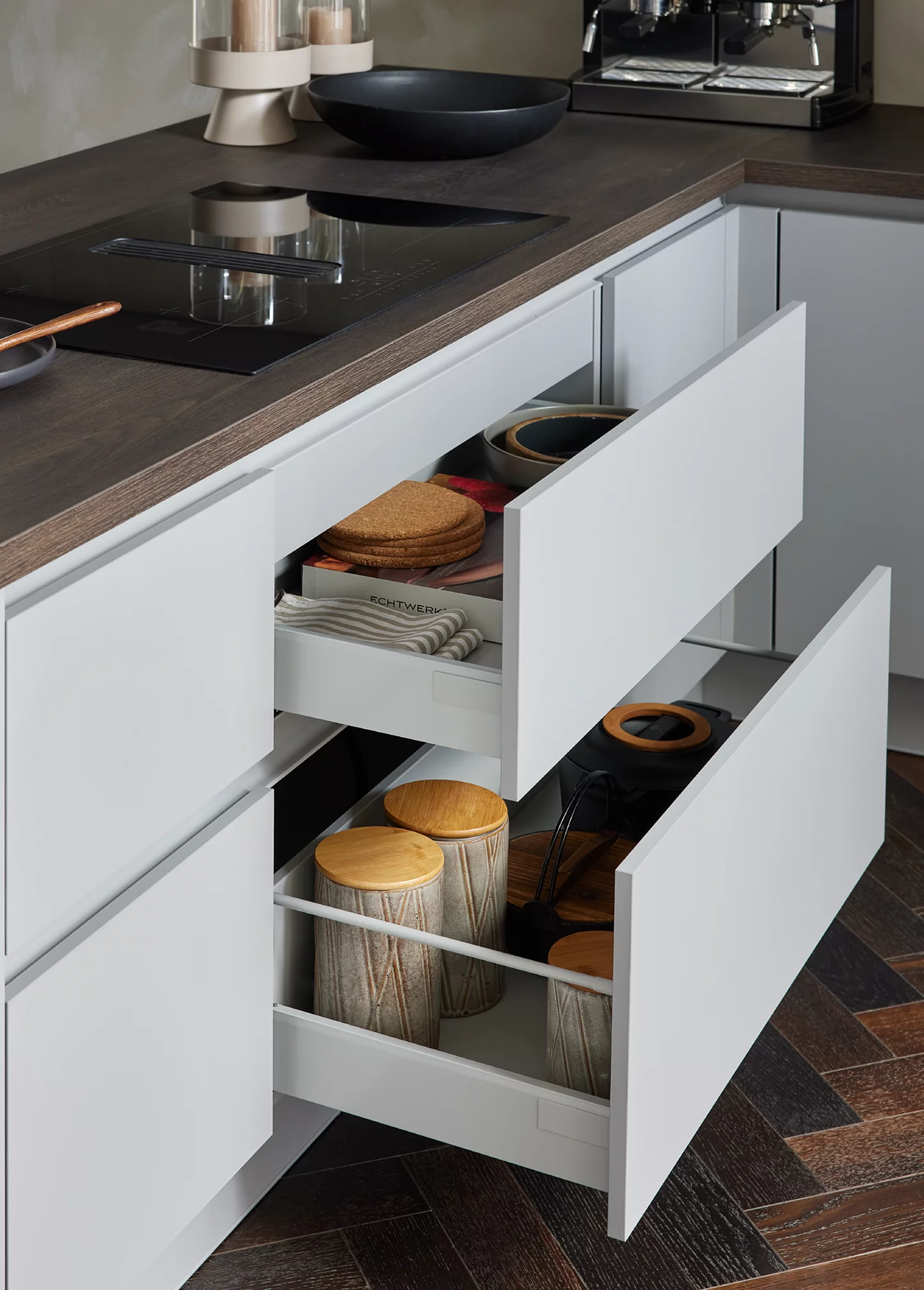 Moderne weiße Küche mit grifflosen, geöffneten Schubladenauszügen für Geschirr und Vorratsbehälter, dunkler Arbeitsplatte und Induktionskochfeld mit Muldenlüfter – funktionale Küchenmöbel mit cleverer Stauraumplanung.